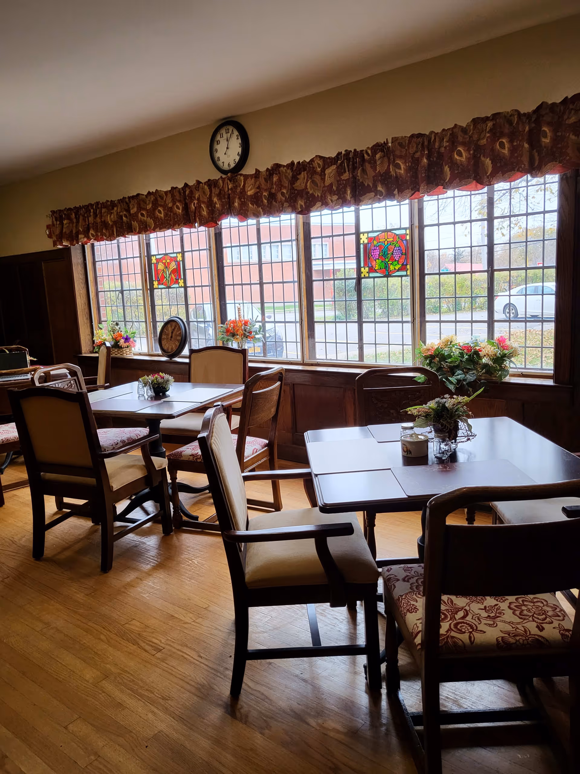 A dining room with wooden floors and several tables and chairs. The tables have placemats and small floral centerpieces. Large windows with decorative stained glass panels and floral valances let in natural light. A clock is mounted on the wall above the windows.