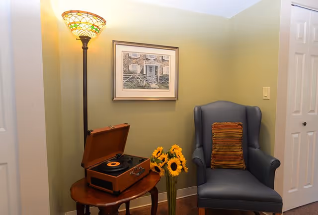A cozy corner with a blue armchair featuring a striped cushion, a wooden side table holding a vintage record player with a vinyl record, a tall floor lamp with a stained glass shade, a vase of sunflowers on the floor, and a framed picture of a house on a light green wall.