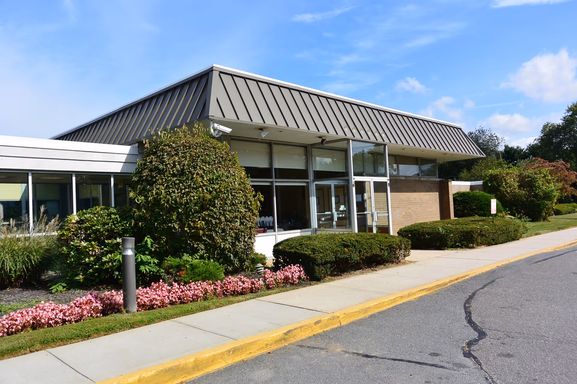 Exterior view of Carillon Nursing and Rehabilitation Center building with a slanted metal roof, large windows, and a sidewalk bordered by neatly trimmed bushes and pink flowers under a blue sky.