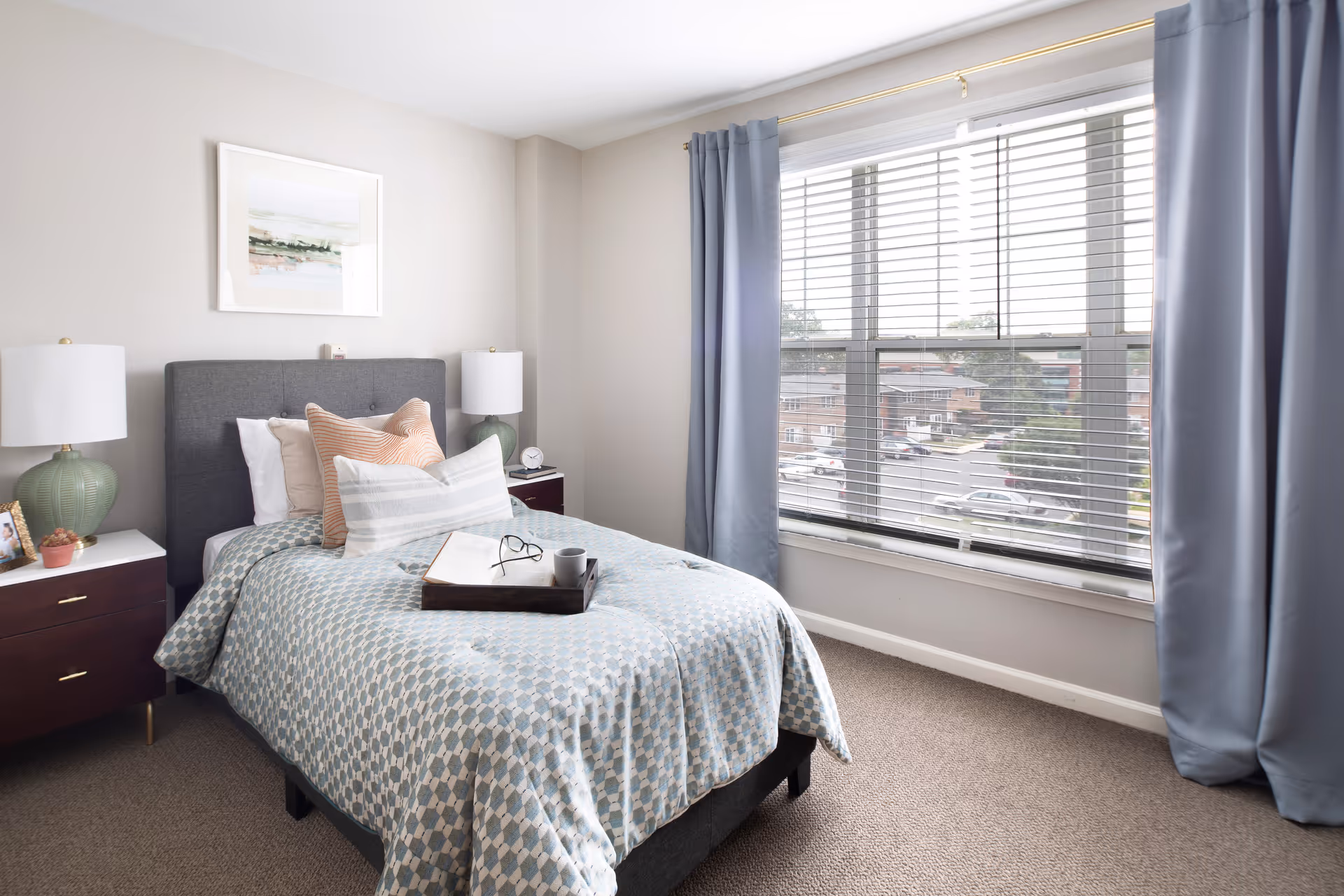 A neatly made single bed with a blue and white patterned bedspread, several pillows, and a tray holding an open book, glasses, and a mug. The room has two bedside tables with matching green lamps, a framed picture on the wall above the bed, and a large window with blue curtains and white blinds overlooking a parking lot and buildings outside.