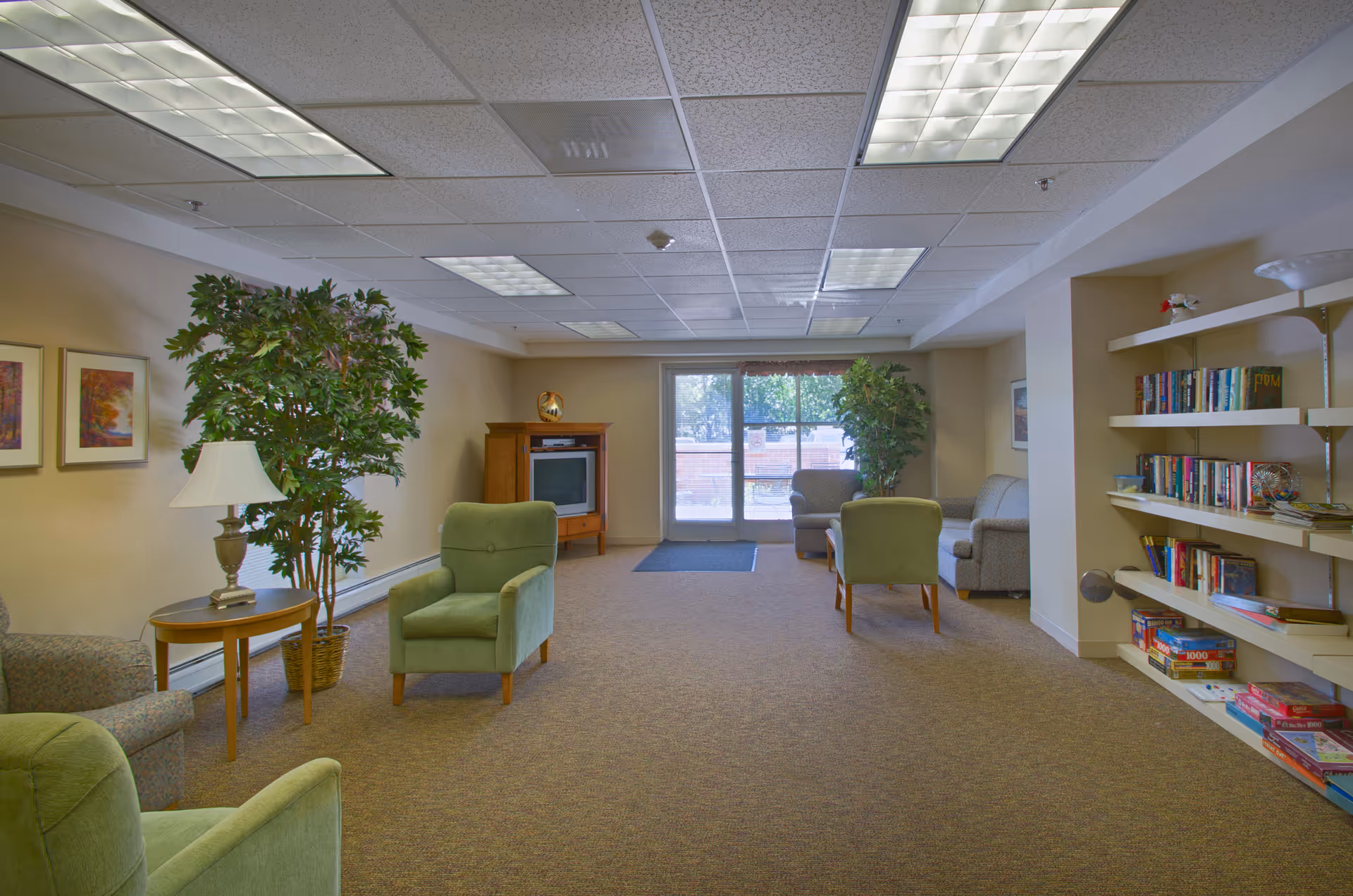A senior living common lounge with green armchairs, a TV cabinet, bookshelves, potted plants, and glass doors to the outside.