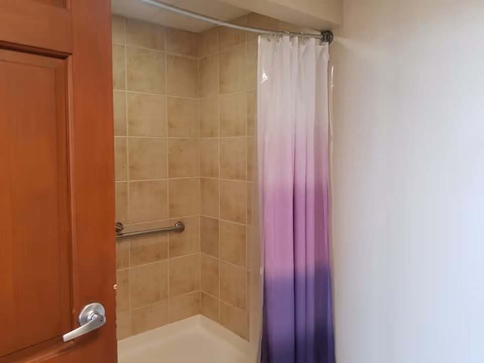 A shower area with beige tiled walls and a white shower base. There is a metal grab bar attached to the wall and a shower curtain with a gradient color from white at the top to purple at the bottom. A wooden door with a silver handle is partially open to the left.