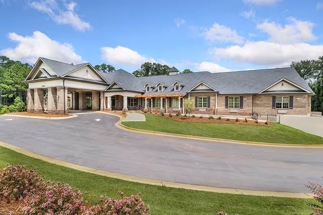 Front exterior of a single-story brick senior living facility with a covered drive and landscaped lawn under a blue sky.