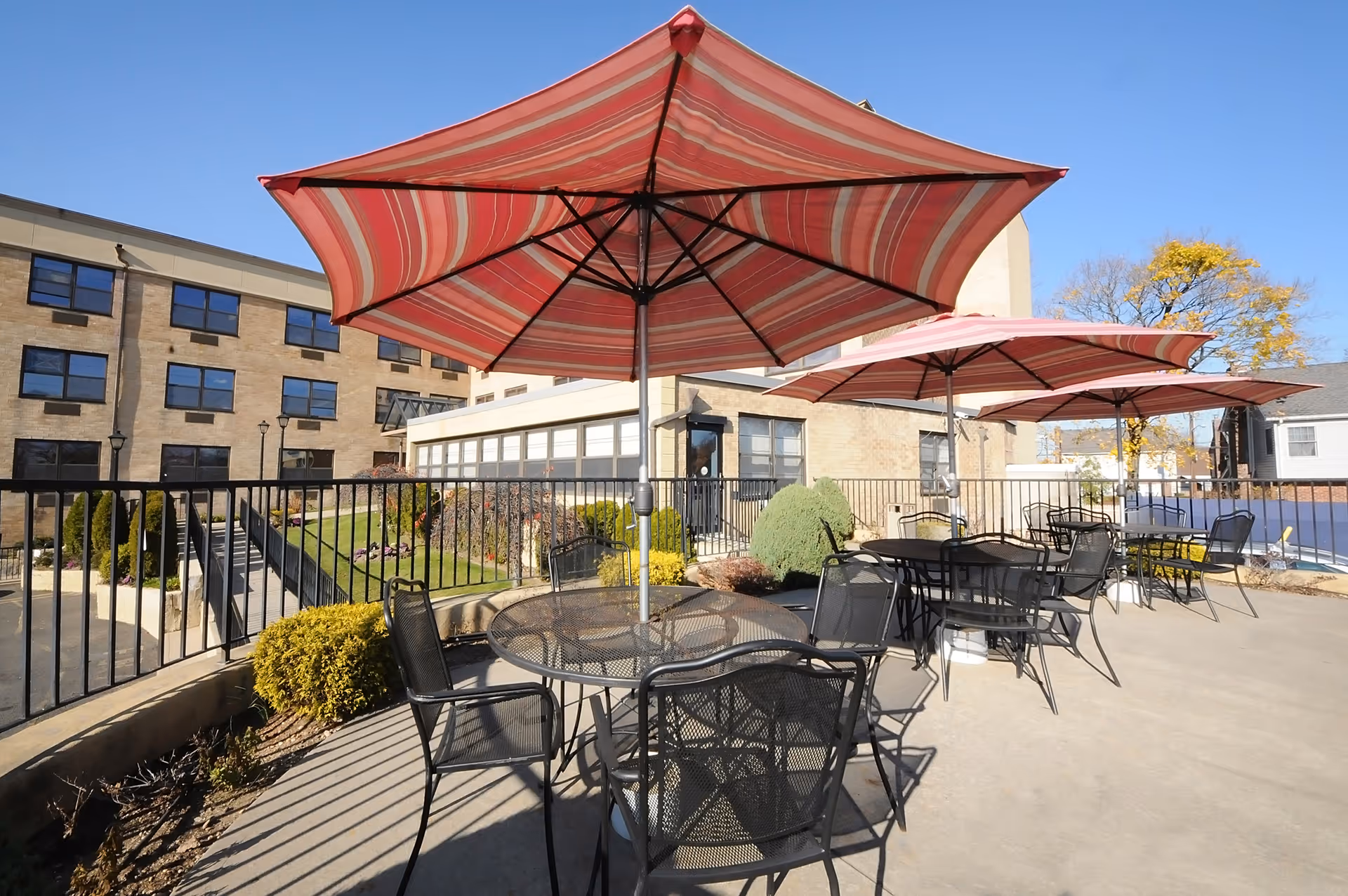 Outdoor patio area at Garden Care Center with several black metal tables and chairs under large red and white striped umbrellas. The patio is surrounded by a black metal fence and there is a multi-story brick building in the background under a clear blue sky.