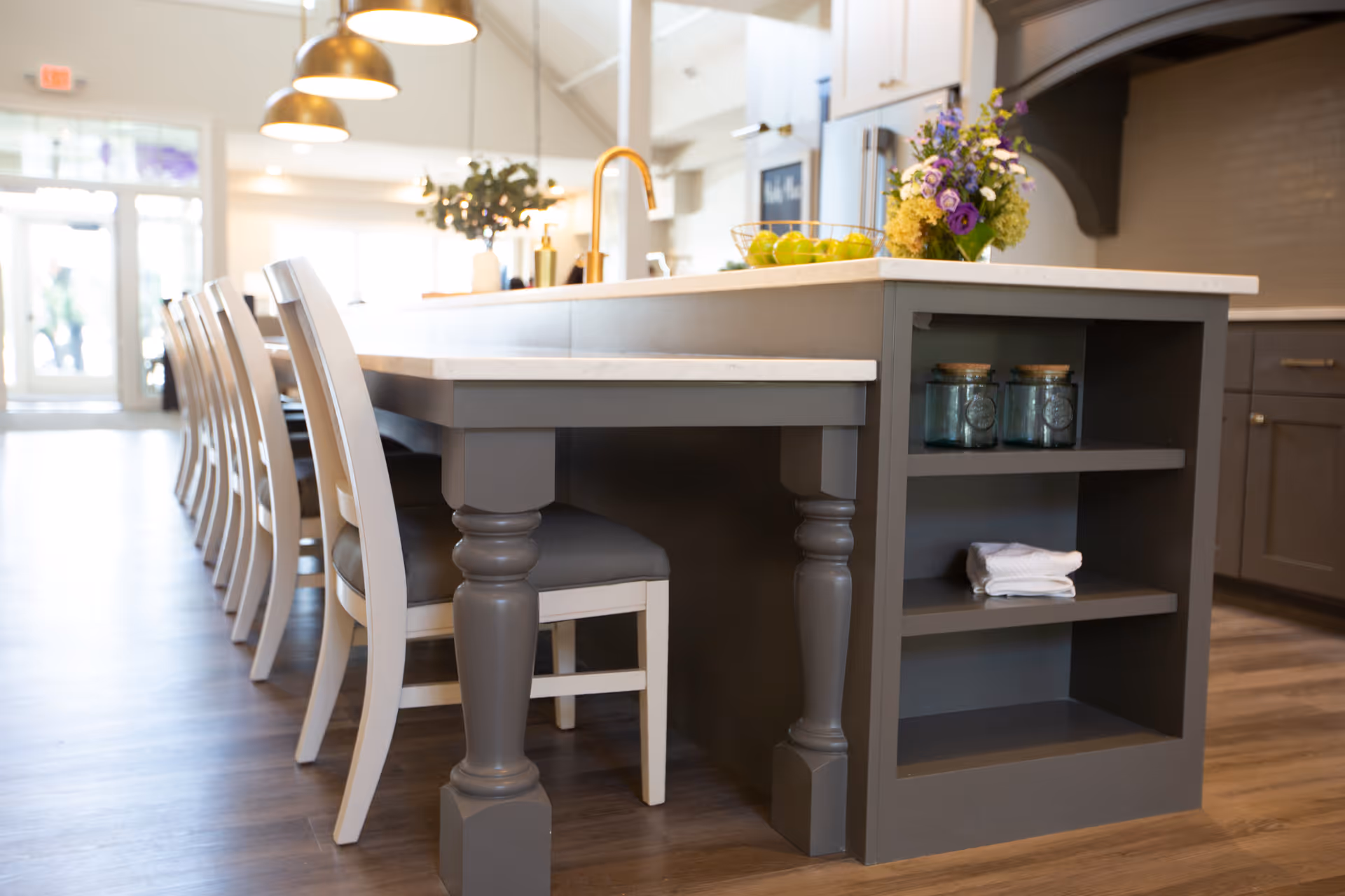 A modern kitchen island with a white countertop and gray base, featuring built-in shelves holding glass jars and a folded towel. Several white chairs with gray cushions are lined up along one side of the island. The kitchen has wooden flooring, pendant lights hanging above the island, and a vase with flowers and a bowl of green apples on the countertop. The background shows a bright entrance with large windows.