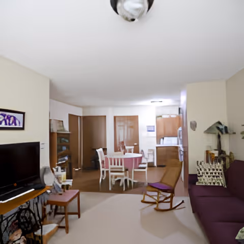 Open living room with a purple sofa, TV and rocking chair, with a dining table and kitchen visible in the background.