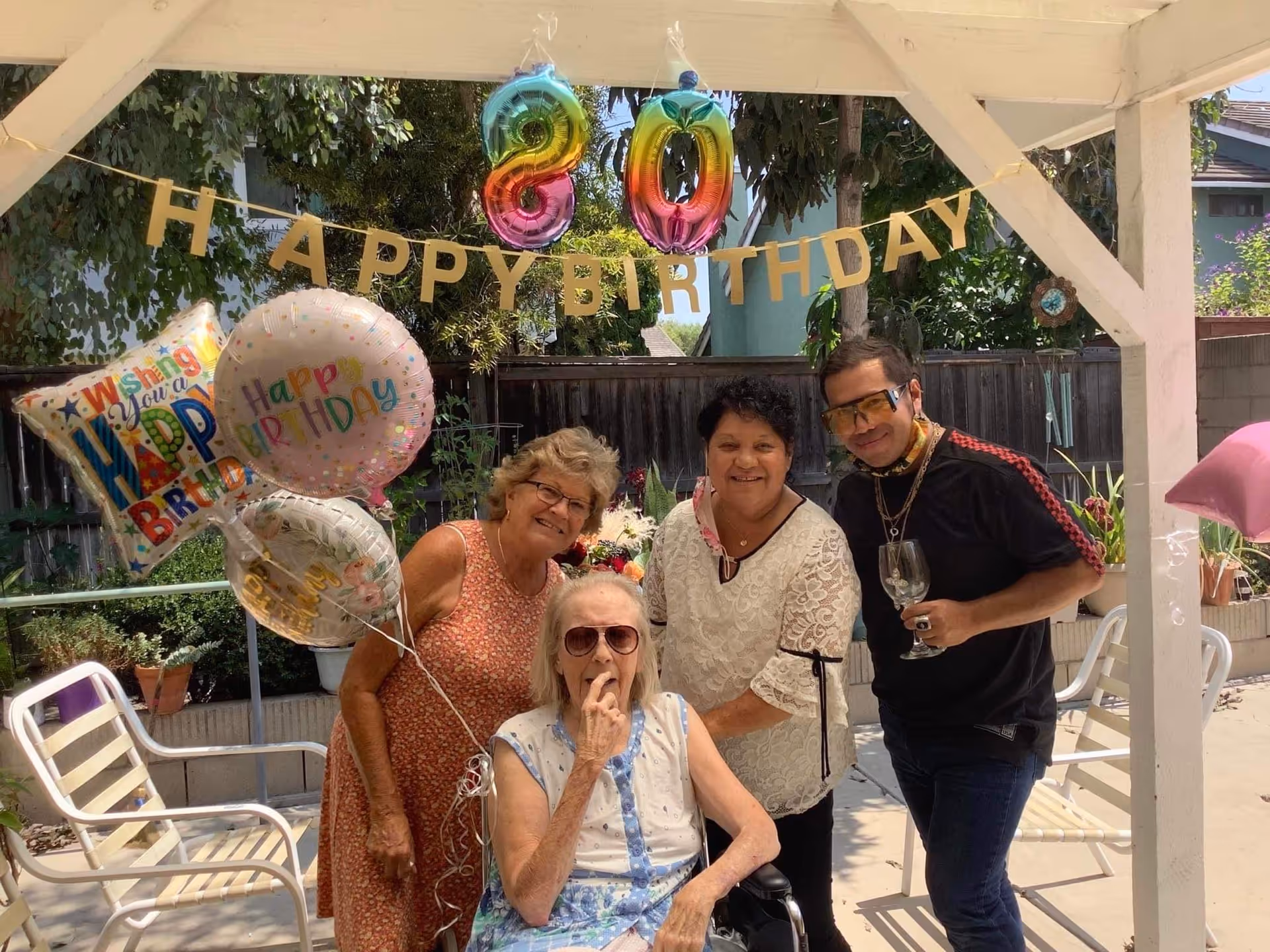 Four people celebrating a birthday outdoors under a white pergola. There are colorful balloons and a 'HAPPY BIRTHDAY' banner hanging above them. One elderly woman is seated in a wheelchair wearing sunglasses, while three other adults stand behind her smiling. The setting appears to be a garden or patio area with greenery and outdoor furniture.