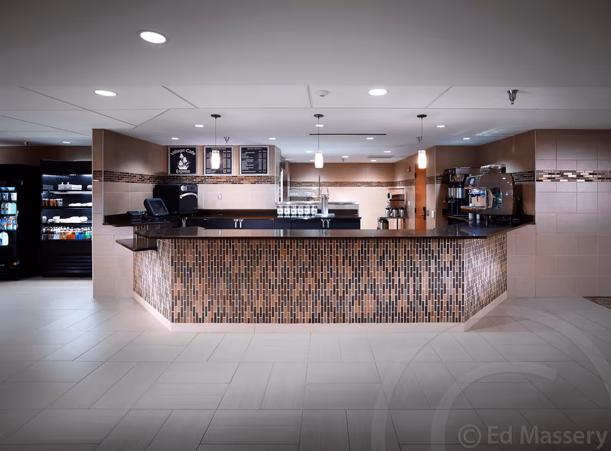 A modern cafe counter area with a tiled front and dark countertop, featuring coffee machines, a menu board, and refrigerated display cases with beverages and snacks in the background.