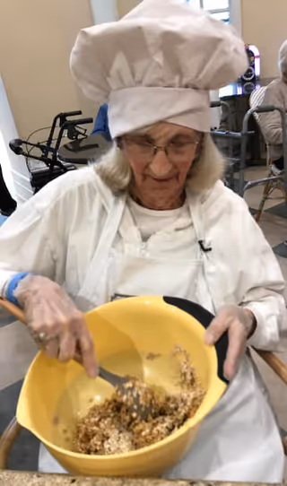 An elderly woman wearing a white chef's hat and apron is mixing ingredients in a large yellow bowl while seated indoors. There are walkers and other people in the background.