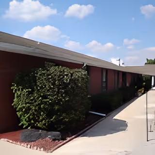 Exterior view of a single-story building with a sloped roof, red walls, and large green bushes along the side. The sky is blue with scattered clouds.