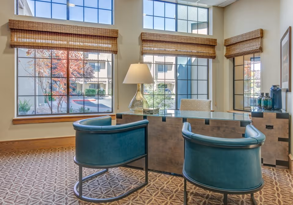 A bright office space with a glass-top desk, two teal upholstered chairs, a table lamp, and large windows with bamboo blinds showing an exterior view of a courtyard with trees and buildings.