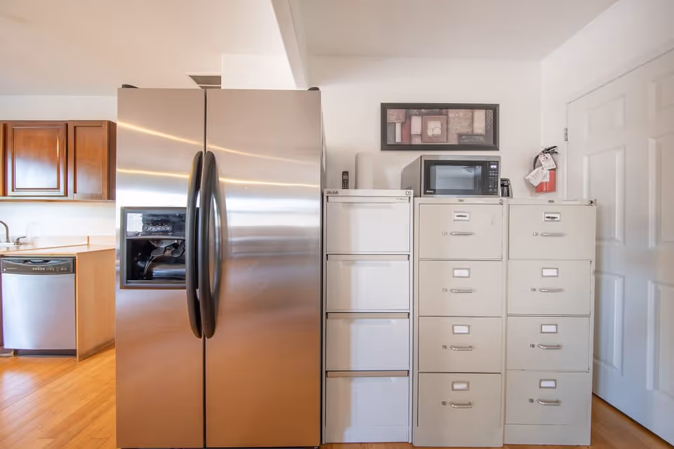 Stainless steel side-by-side refrigerator and dishwasher in a kitchen area next to stacked filing cabinets topped with a microwave and a fire extinguisher.