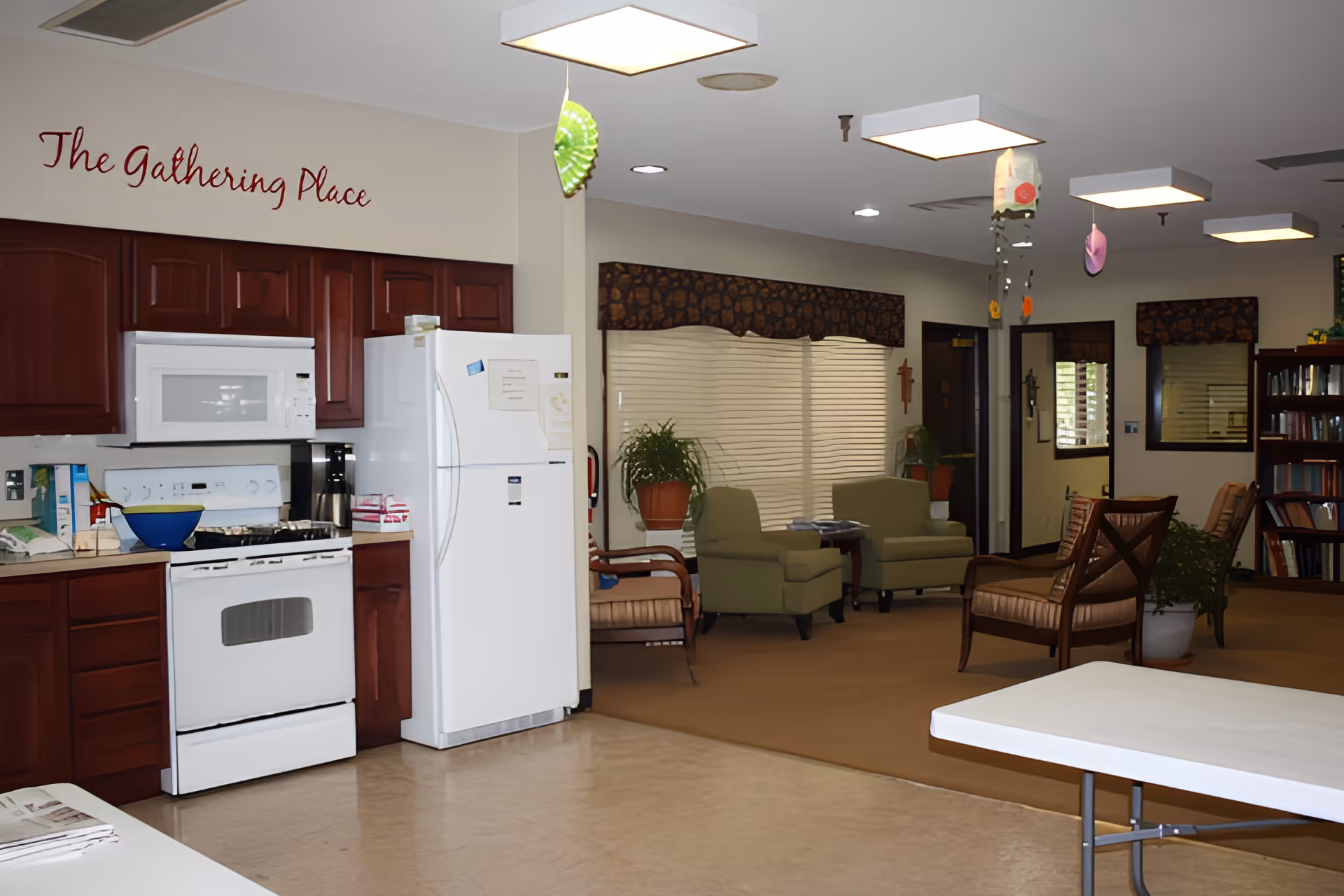 Communal lounge and kitchenette with a stove and refrigerator on the left and armchairs and tables on the right beneath a sign reading 'The Gathering Place'.