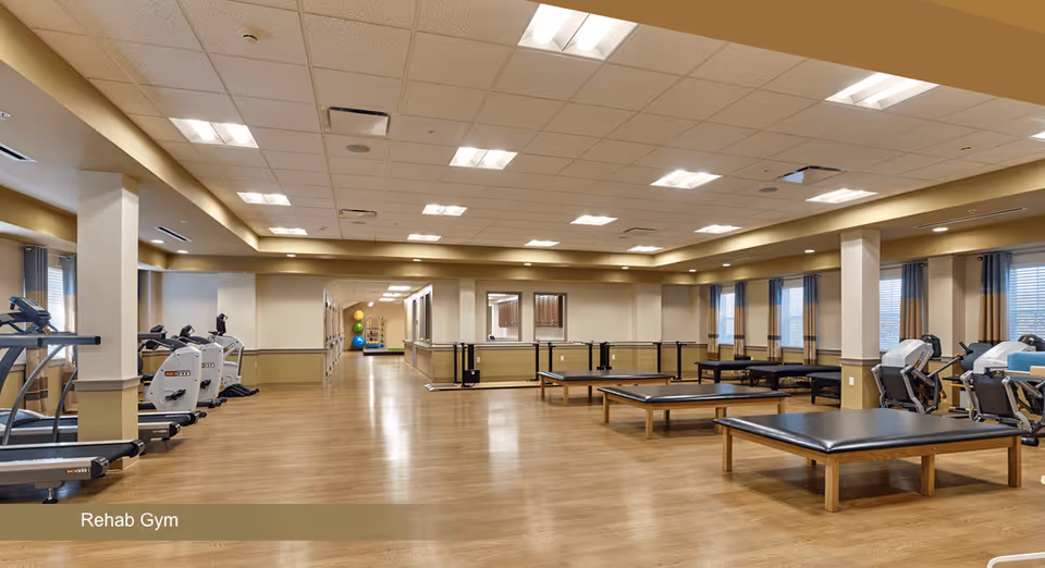 A spacious rehab gym with wooden flooring, multiple exercise machines including treadmills and stationary bikes on the left side, and several padded therapy tables on the right. The room has large windows with curtains, bright ceiling lights, and a hallway leading to another area in the background.