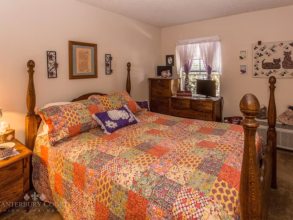 A cozy bedroom with a wooden four-poster bed covered in a colorful patchwork quilt. There are matching pillows on the bed, a wooden nightstand with a lamp and tissue box, and a wooden dresser with a small TV and decorative items. The window has white curtains with purple accents, and the walls are decorated with framed art and quilted wall hangings.