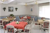 A bright dining room with several tables covered in red and beige tablecloths, each decorated with small floral centerpieces. The chairs have floral upholstery. The walls are light-colored with a blue wainscoting and decorated with framed pictures and wall-mounted plants. Large windows with blinds allow natural light to fill the room.