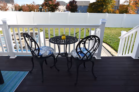 A small outdoor patio area with a black metal table and two matching chairs with blue and white cushions. The patio has dark wooden flooring and white railing. Beyond the patio is a green lawn enclosed by a white fence, with houses and trees showing autumn foliage in the background.