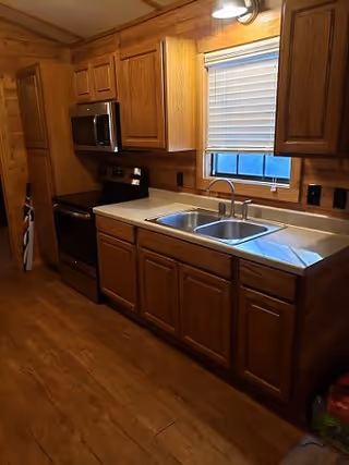 A kitchen with wooden cabinets and drawers, a double stainless steel sink under a window with closed blinds, a black stove with an oven, and a microwave above it. The floor is wooden, and the walls have a wood panel finish.