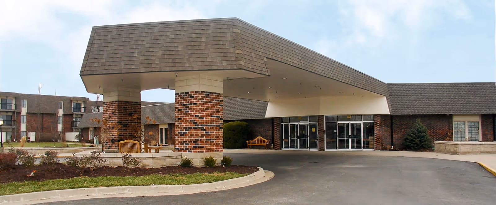 Exterior view of Foxwood Springs Senior Living facility showing the main entrance with a covered drop-off area supported by brick columns, benches near the entrance, and a multi-story building in the background.