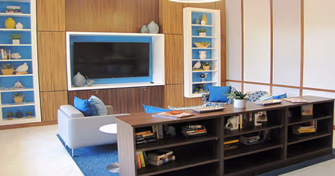 A modern living room area featuring a large flat-screen TV mounted on a wooden wall with blue and white shelving units on either side displaying decorative items. In front of the TV, there is a gray armchair with blue cushions and a wooden bookshelf filled with books and magazines. A blue rug is placed under the armchair and bookshelf, and a small white round table is next to the armchair.