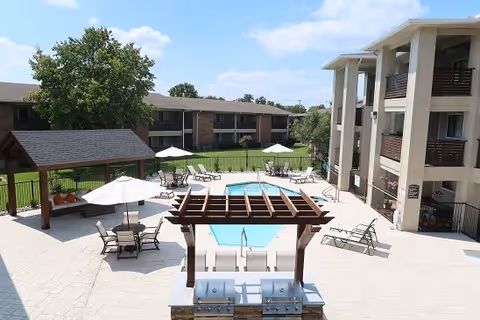 Outdoor pool area at MontClair Estates featuring a swimming pool surrounded by lounge chairs and tables with umbrellas. There is a wooden pergola with two grills underneath and a shaded seating area with pumpkins on a bench. The area is enclosed by a fence with apartment buildings and trees in the background under a blue sky with some clouds.