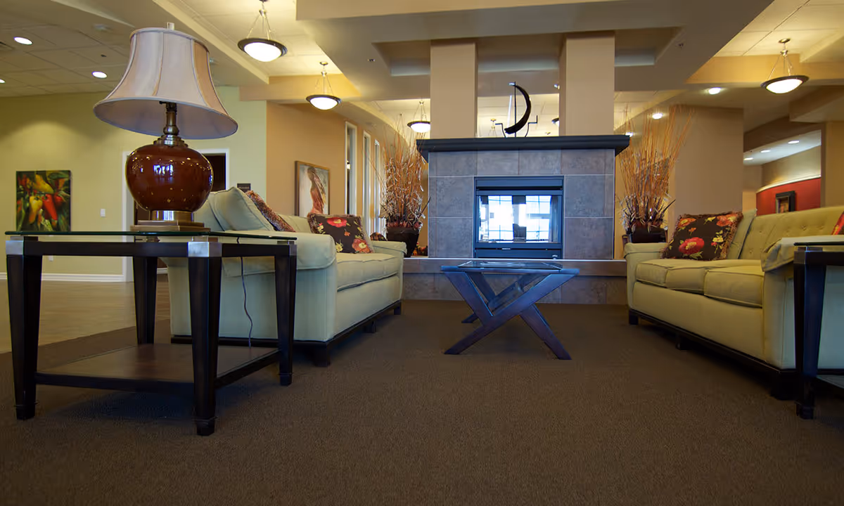 A cozy senior living facility common area with two light green sofas facing each other, a glass coffee table in the center, and a fireplace with a tiled surround in the background. There are decorative plants and framed artwork on the walls, with warm lighting from ceiling fixtures.