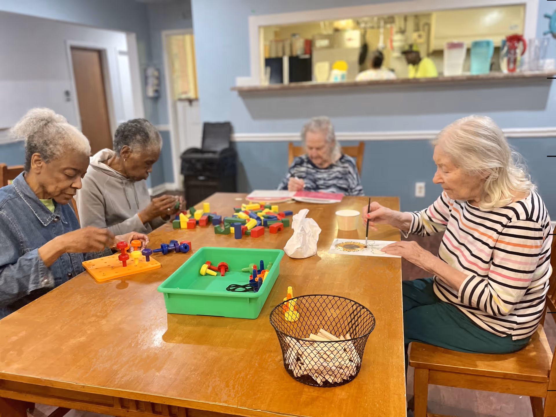 Four elderly women sitting around a wooden table engaged in various activities including painting and playing with colorful pegboards and blocks in a room with light blue walls and a serving window to a kitchen area.