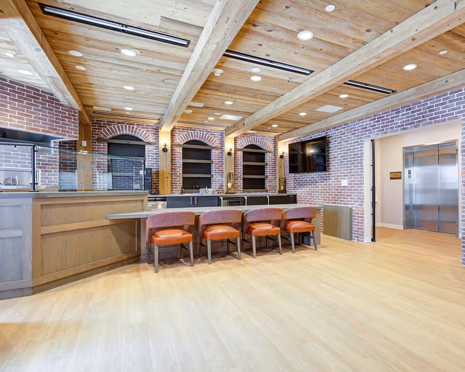 Interior view of a common area with a wooden ceiling featuring exposed beams and recessed lighting. The room has brick walls with three arched alcoves containing shelves and a counter with a sink. In front of the counter are four orange cushioned chairs. A flat-screen TV is mounted on the wall to the right, and an elevator is visible through an open doorway on the far right.