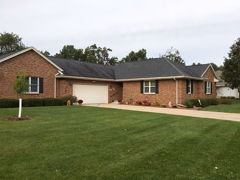 Exterior view of a single-story brick building with a dark shingled roof, a two-car garage, and a well-maintained green lawn with a small tree and flower beds near the entrance.
