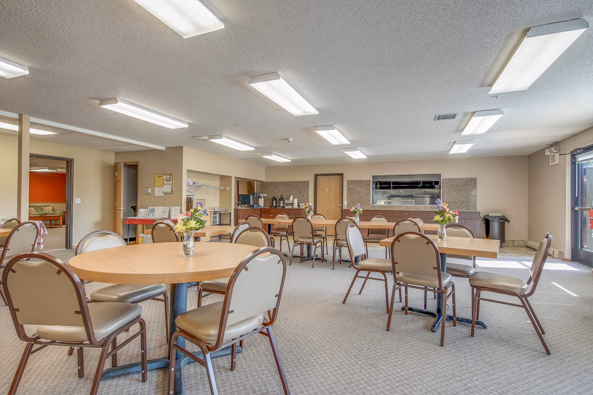 Communal dining room with round tables and chairs, a serving counter, and vases of flowers under fluorescent ceiling lights.