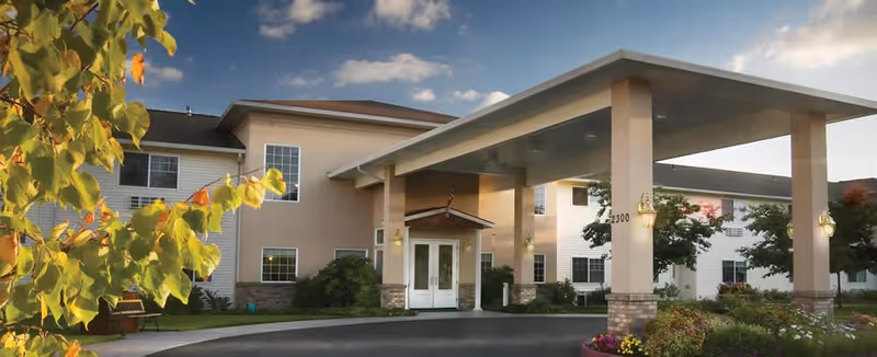 Exterior view of Columbia Ridge Senior Living facility showing a two-story building with beige and white walls, multiple windows, a covered entrance with pillars, and landscaped greenery including trees and flowers under a partly cloudy sky.