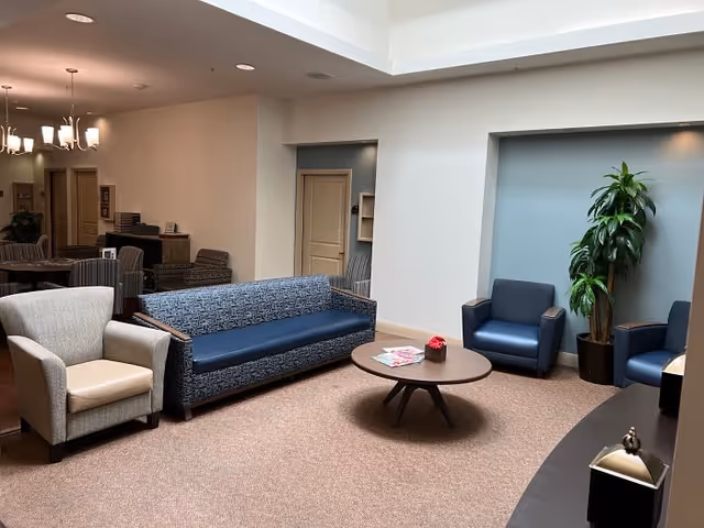 Communal seating area with sofas, armchairs, a round coffee table, and a potted plant in a senior living facility.