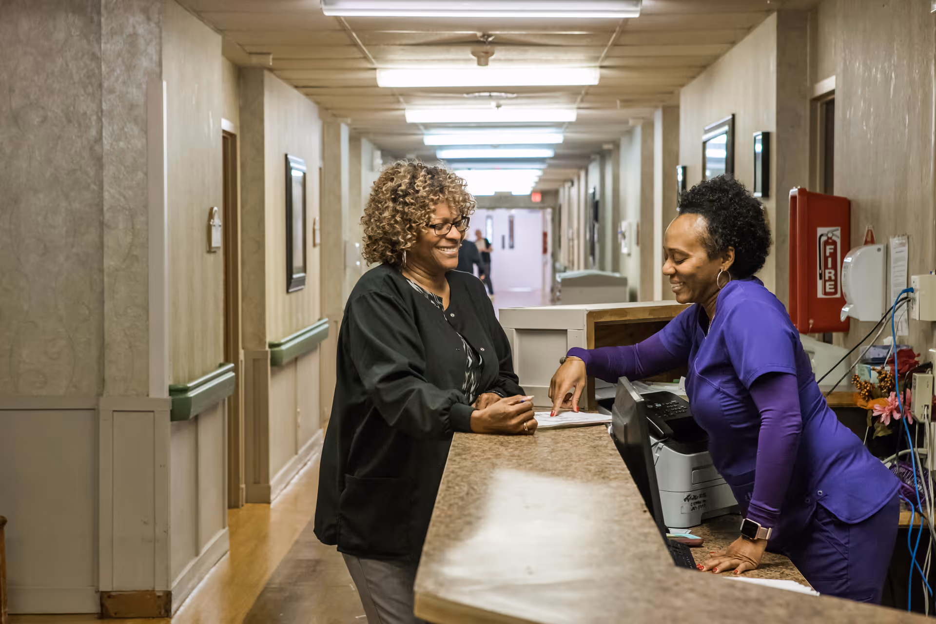Two women smiling and talking at a reception desk in a hallway of a senior living facility. One woman is standing behind the desk wearing purple scrubs, and the other woman is standing in front of the desk wearing a black jacket. The hallway has several doors and framed pictures on the walls.