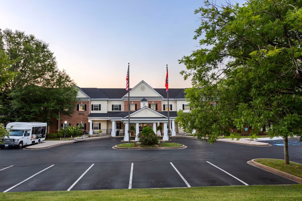 Front exterior view of StoryPoint Chattanooga building with two flagpoles displaying the American and Tennessee state flags, a covered entrance, a parking lot with marked spaces, a shuttle bus on the left, and trees on both sides.