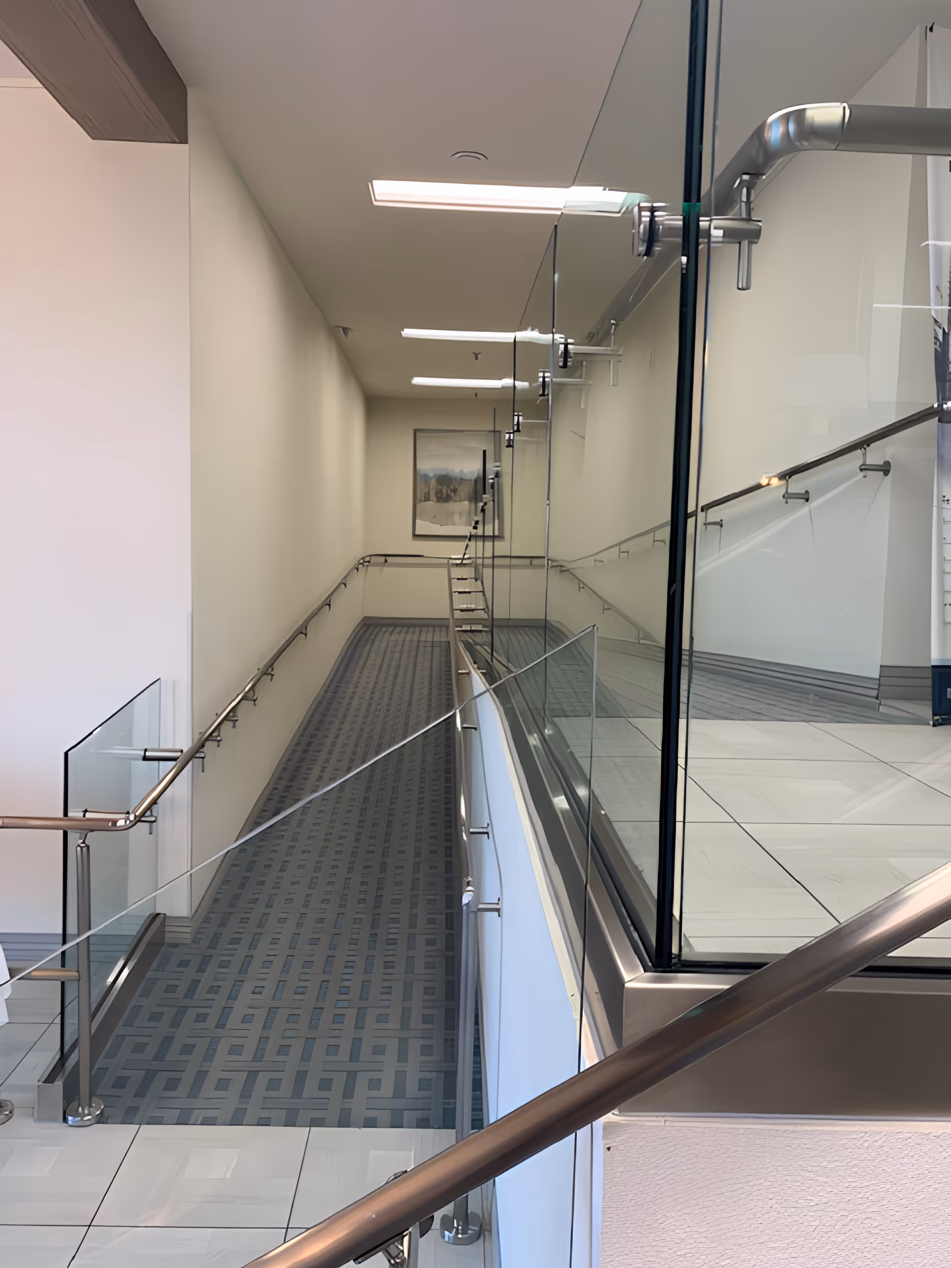 Interior view of a modern hallway with a long, gently sloping ramp covered in patterned carpet. The ramp is flanked by stainless steel handrails and glass panels. The walls are white, and there is a framed picture at the end of the hallway. The ceiling has recessed lighting fixtures.