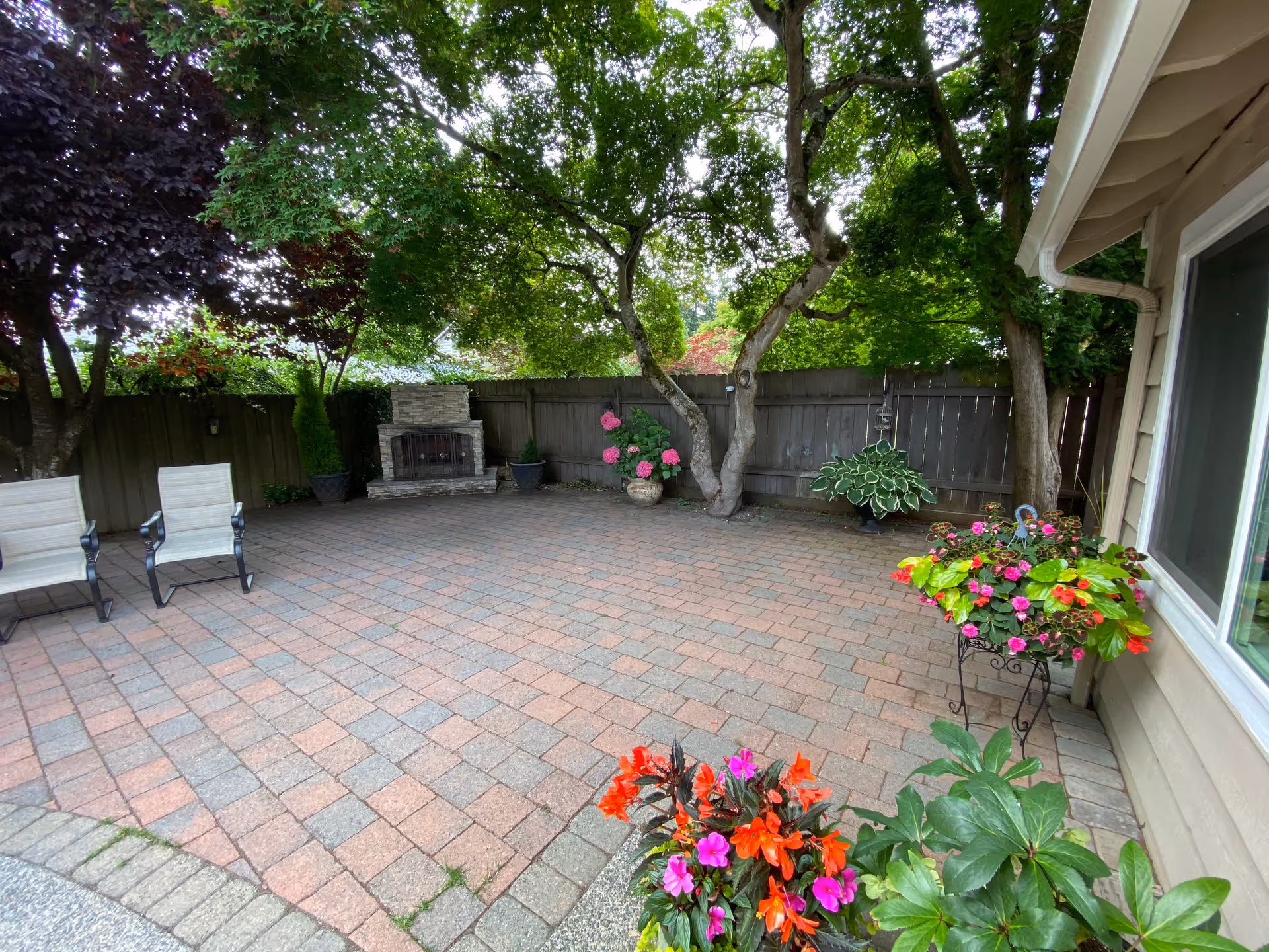 A peaceful outdoor patio area with brick pavers, two white chairs, a stone fireplace, and various potted plants and flowers. The space is surrounded by a wooden fence and shaded by large trees.