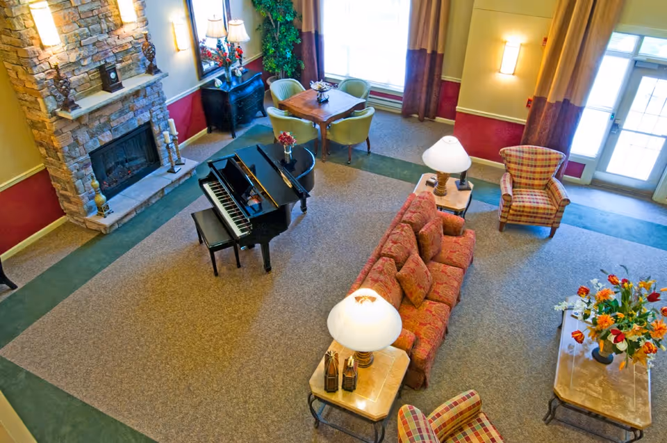 A cozy senior living facility common area with a stone fireplace, a black grand piano, a red patterned sofa, two plaid armchairs, side tables with lamps, a wooden table with four chairs near a large window, and a coffee table with a colorful flower arrangement.