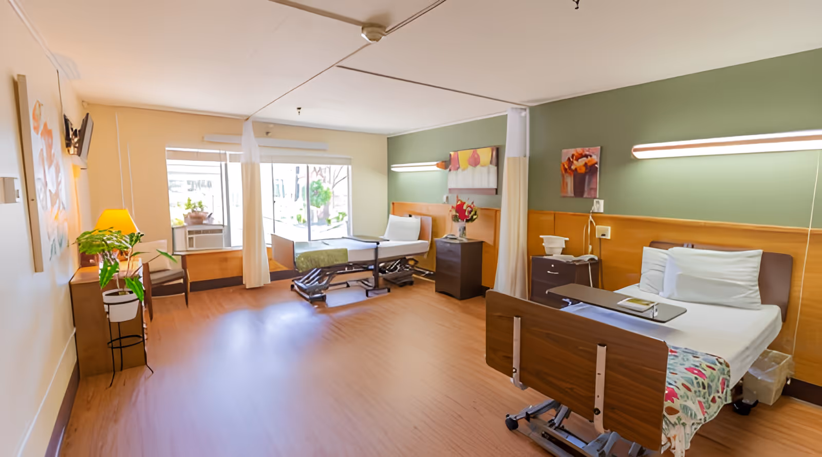 A bright shared nursing facility room with two hospital beds, bedside tables, chairs, plants, and large windows.