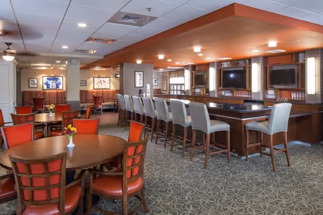 Spacious dining area with round wooden tables and red cushioned chairs, a long bar counter with high chairs, and framed sports memorabilia on the walls. The room has a patterned carpet and recessed lighting in the ceiling.