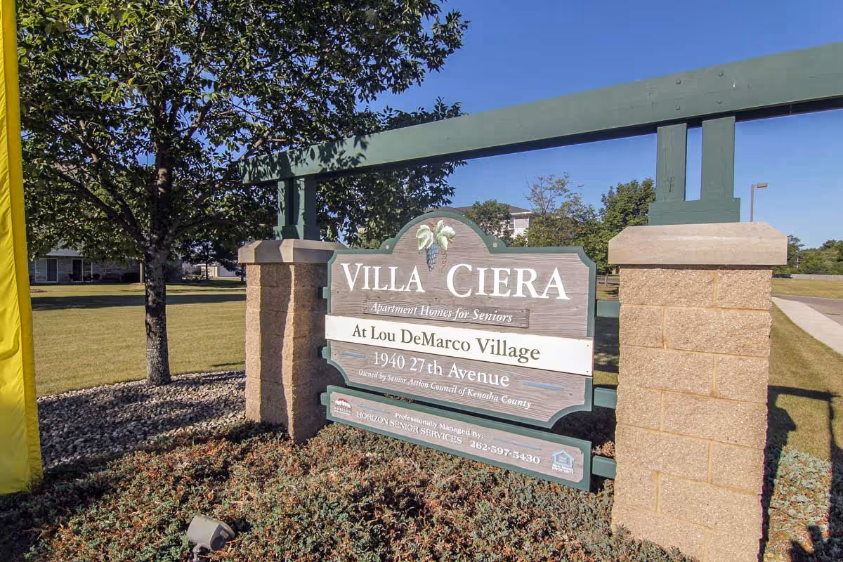Outdoor view of a large wooden sign for Villa Ciera Apartment Homes for Seniors at Lou DeMarco Village, located at 1940 27th Avenue, surrounded by grass, trees, and a clear blue sky.