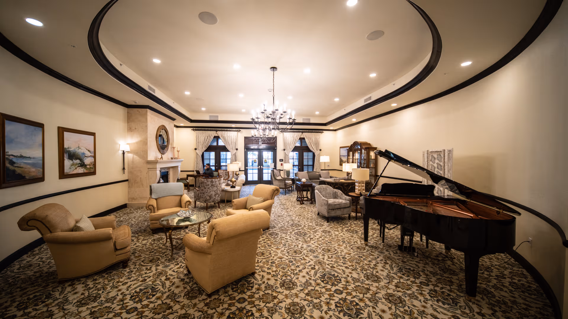 Spacious and elegant living room area with patterned carpet, beige and gray upholstered armchairs, a grand piano on the right, a chandelier hanging from the ceiling, and a fireplace on the left wall. The room has large windows with curtains and framed paintings on the walls.