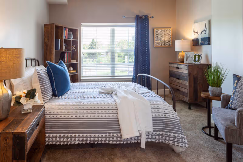 A cozy bedroom with a bed featuring a striped gray and white comforter and blue pillows. There is a wooden nightstand with a lamp and a small flower arrangement on the left side of the bed. A wooden bookshelf with decorative items is next to the window with white blinds and a blue curtain. On the right side, there is a wooden dresser with a lamp, framed photos, and wall art above it. A gray armchair with a pillow and a small round table with a potted plant are also visible.