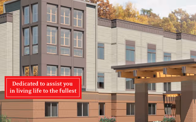 Exterior view of a multi-story assisted living facility building with large windows and a covered entrance. Trees with autumn foliage are visible in the background.