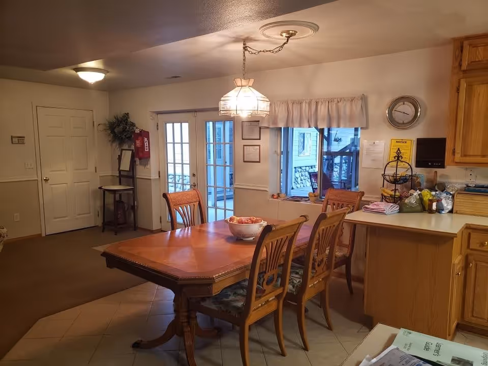 Interior view of a dining area with a wooden dining table and four chairs. A bowl of apples is placed on the table. To the right, there is a kitchen counter with various items on it, including a fruit basket and folded towels. A window with a curtain and a clock on the wall are visible above the counter. French doors and a white door are seen in the background, along with a small table and a fire extinguisher mounted on the wall.