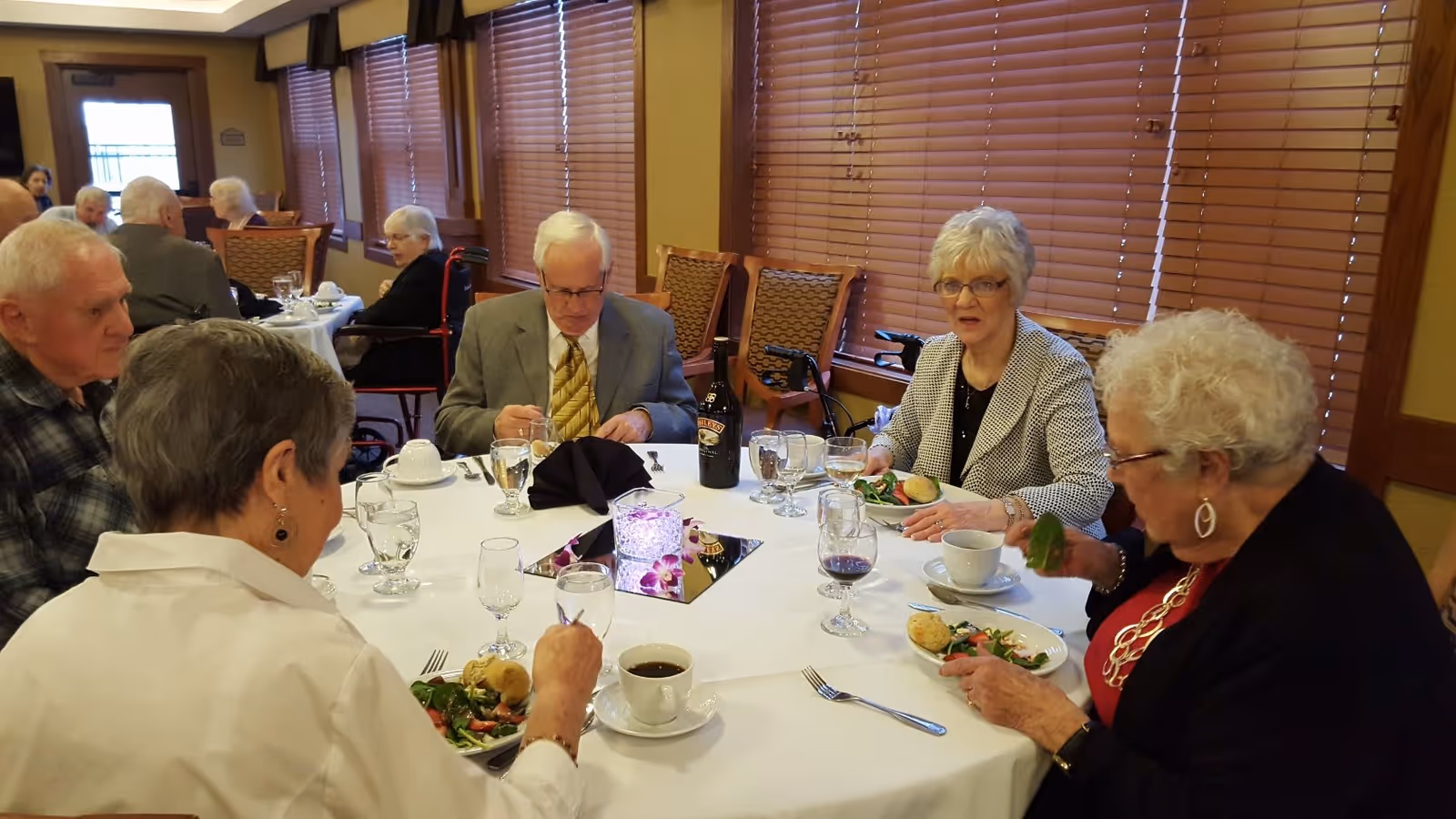 A group of elderly people sitting around a round dining table in a well-lit room with wooden blinds. They are eating a meal that includes salad and bread rolls, with glasses of water, wine, and coffee cups on the table. The atmosphere appears social and communal.