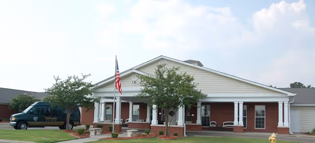 Front exterior view of a single-story brick building with white columns and a covered porch. There are two small trees and an American flag in front of the building, along with a parked van on the left side. The sky is partly cloudy.
