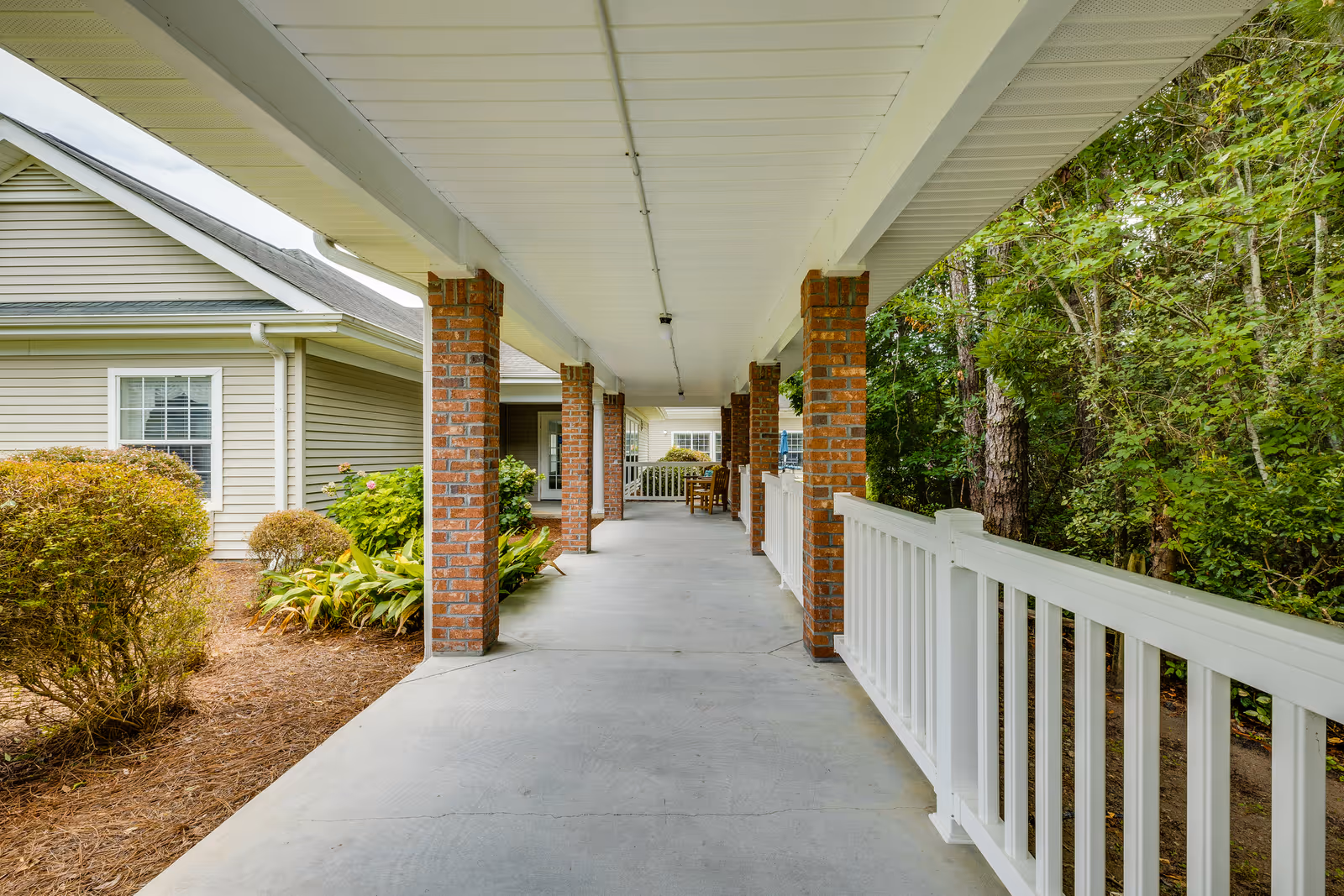 Covered outdoor walkway with brick pillars and white railing, adjacent to a beige building with windows and surrounded by greenery and bushes.