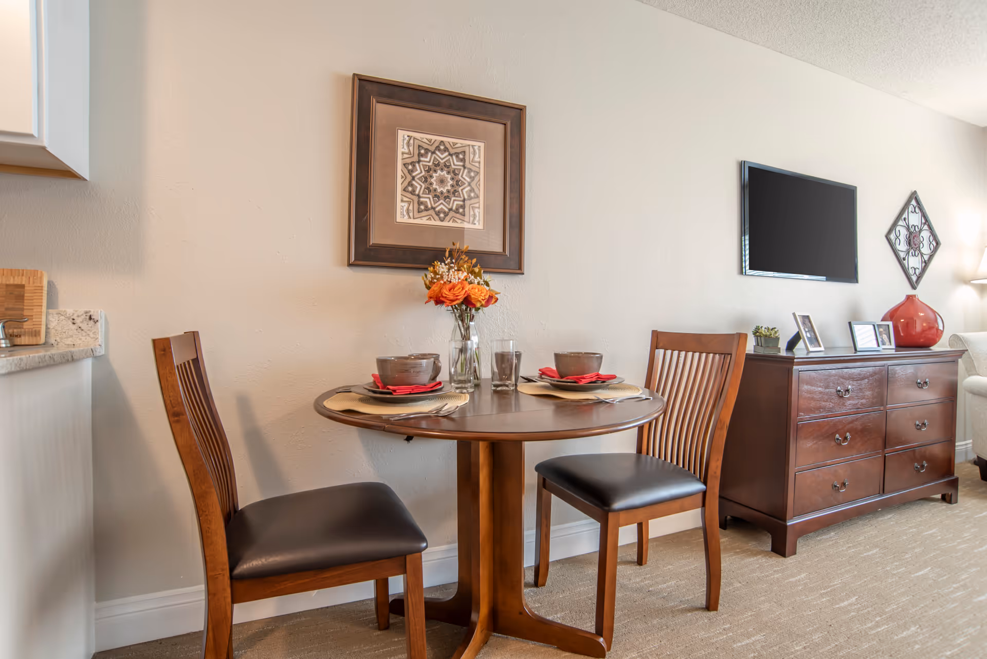 Round wooden dining table set for two with chairs against a wall, framed art above, and a dresser with a TV and decor to the right.