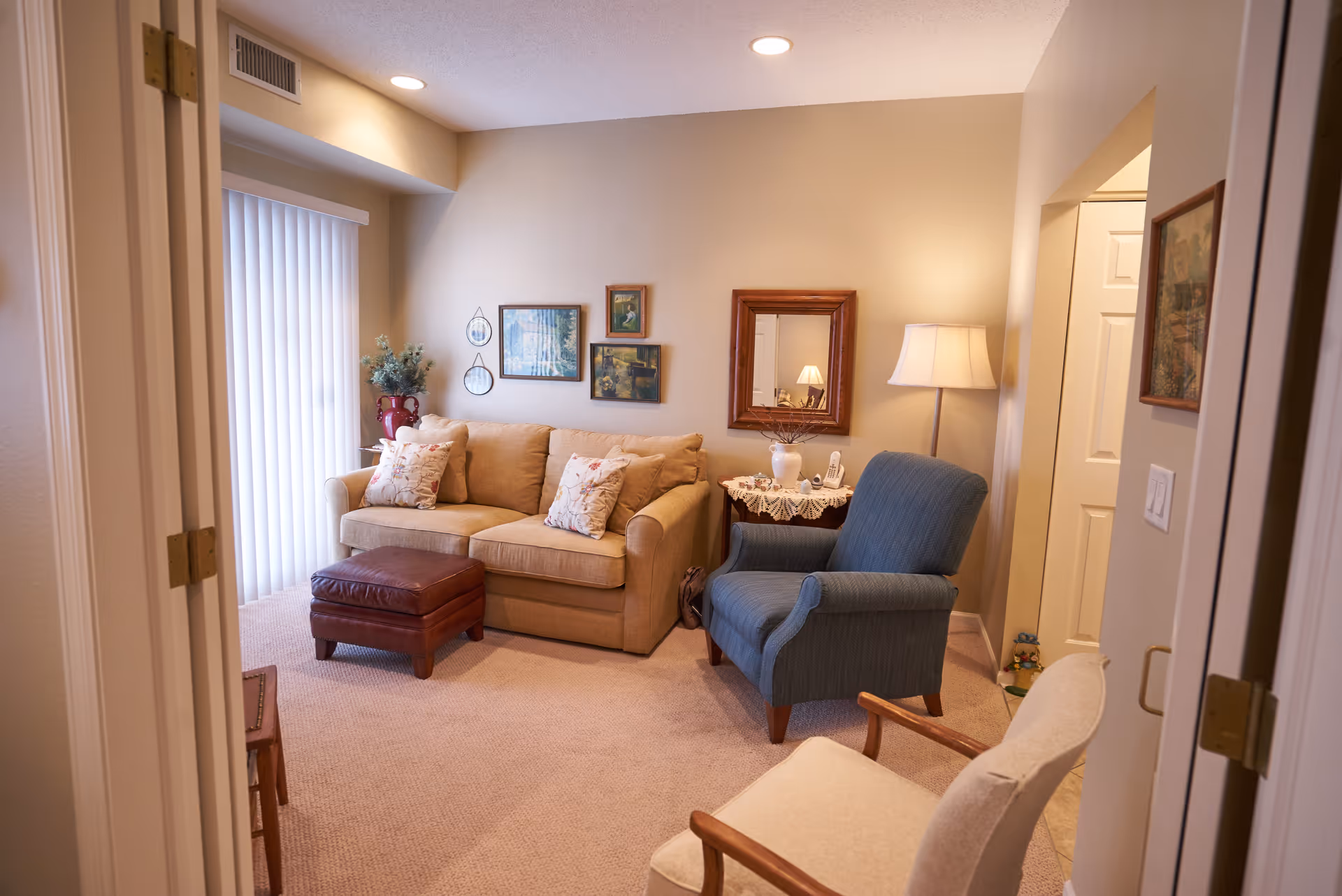 A cozy living room with a beige sofa adorned with floral pillows, a blue armchair, and a beige armchair. There is a brown leather ottoman in front of the sofa. The walls are decorated with framed pictures and a mirror. A side table with a lace doily, a lamp, and a telephone is next to the blue armchair. Vertical blinds cover a window or sliding door on the left side.