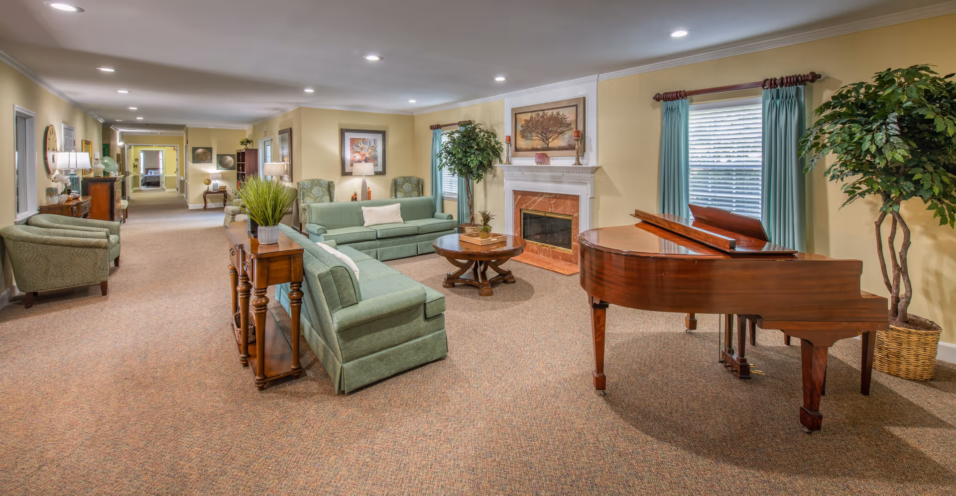A spacious living room area in a senior living facility featuring a grand piano, green upholstered sofas and armchairs, a wooden coffee table, a fireplace with a framed tree artwork above it, large windows with teal curtains, and decorative plants. The room has beige walls and carpeted flooring, with additional seating and furniture visible down a hallway.